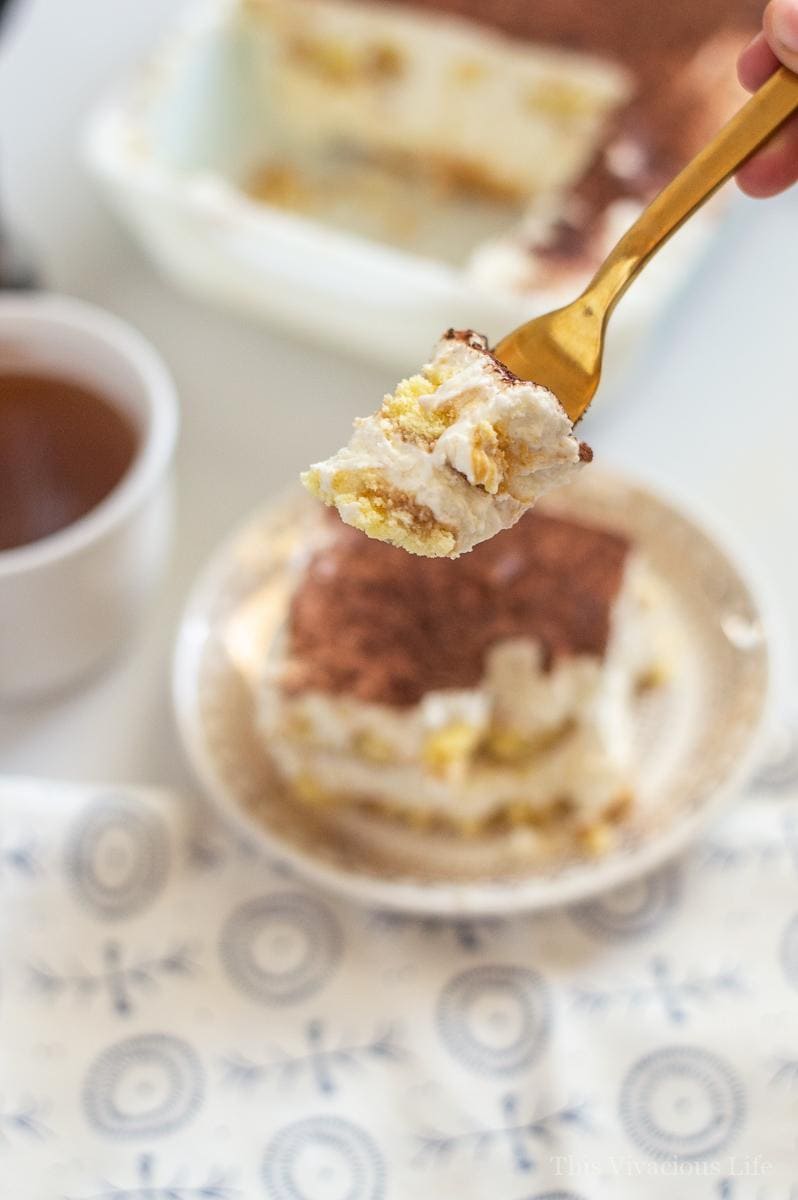 A bite of gluten-free tiramisu on a gold spoon with the tiramisu and cocoa drink in the background. The bite shows the layers of the dessert.