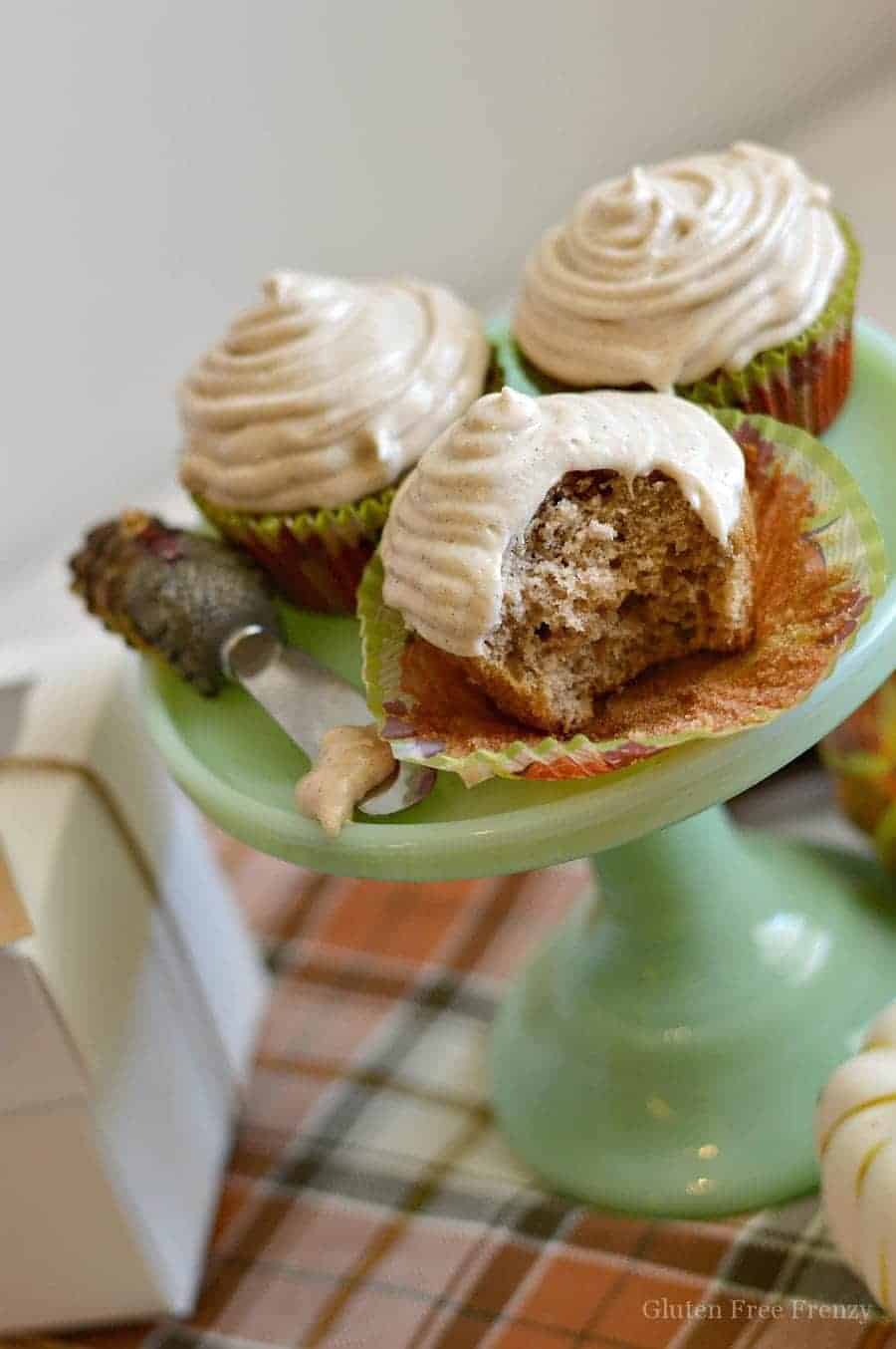 Three frosted apple cider cupcakes on a green cake stand. One cupcake is bitten into so you can see the fluffy texture of the cupcake.