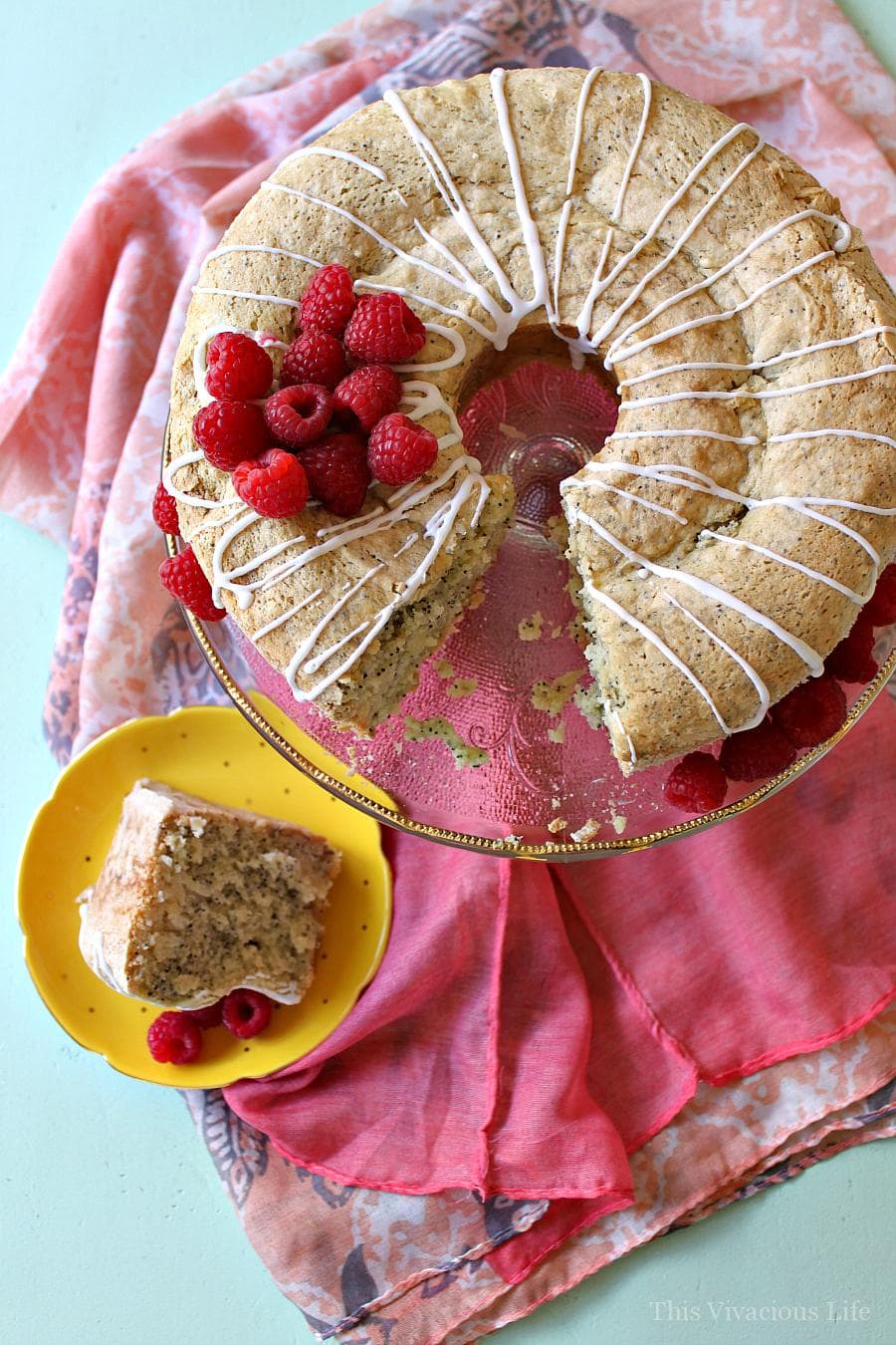 Gluten-free lemon poppy seed bundt cake on cake pan, with a slice cut out and put on a plate. There is a white drizzle glaze and raspberries on top.