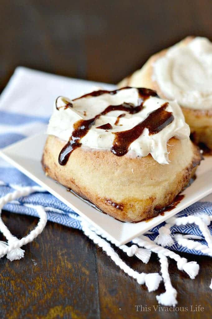 A gluten-free cinnamon roll topped with cream cheese frosting and brown sugar drizzle on a white plate on a blue and white towel.