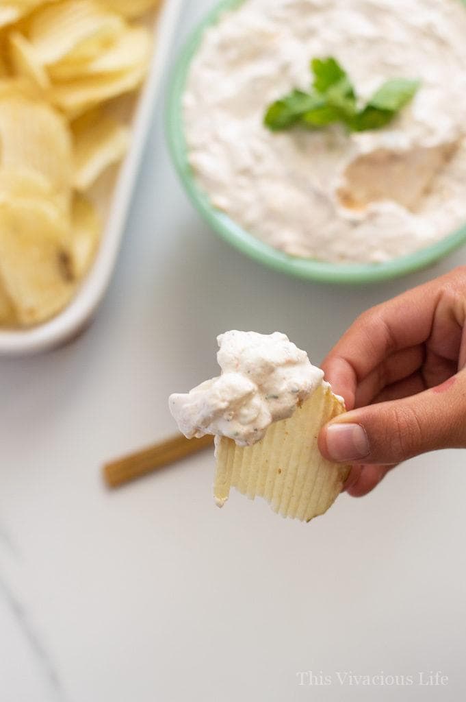 A hand holding a ridged chip covered in Sour Cream Chip Dip with Dip in the background