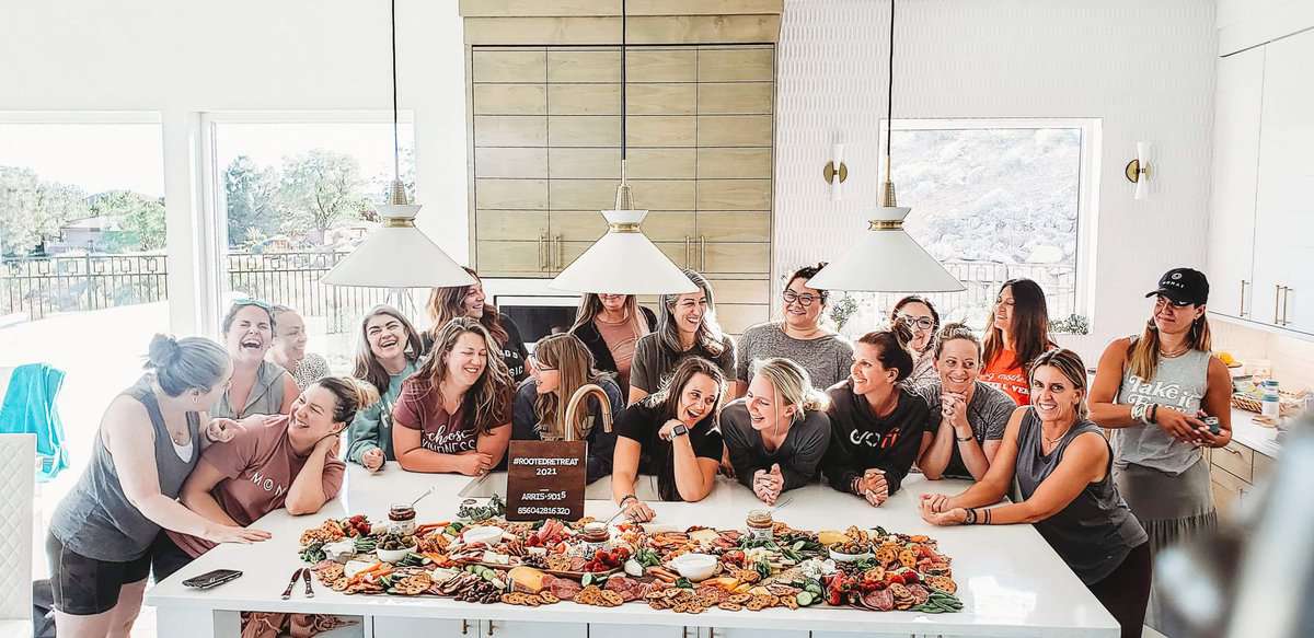 A group of women laughing and sitting around a large charcuterie grazing table filled with meats, cheeses, pretzels, and other snacks