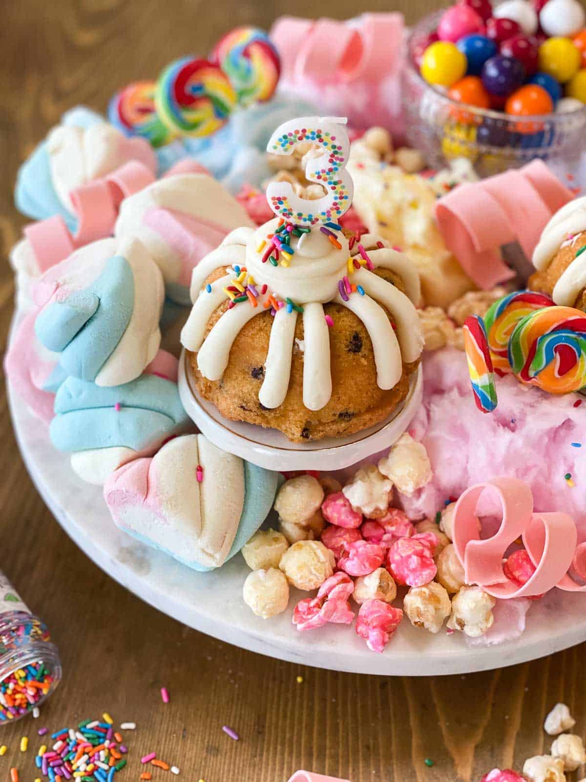 Birthday charcuterie board on a white marble platter. Small bundt cake with white frosting and number 3 on top surrounded by popcorn, gum, gumballs, and cotton candy.