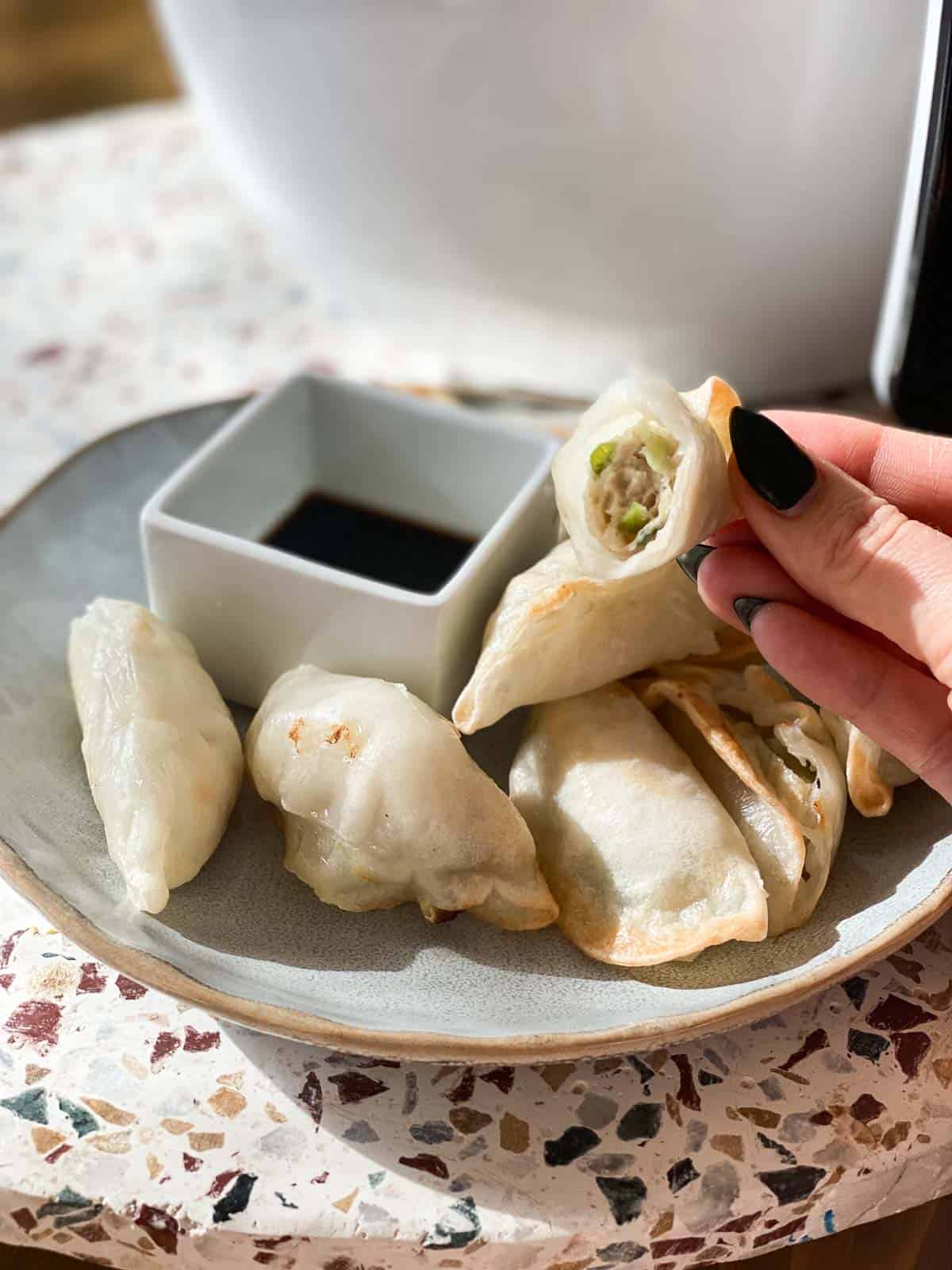 Hand holding partially eaten potsticker with plate of potstickers and sauce in the background