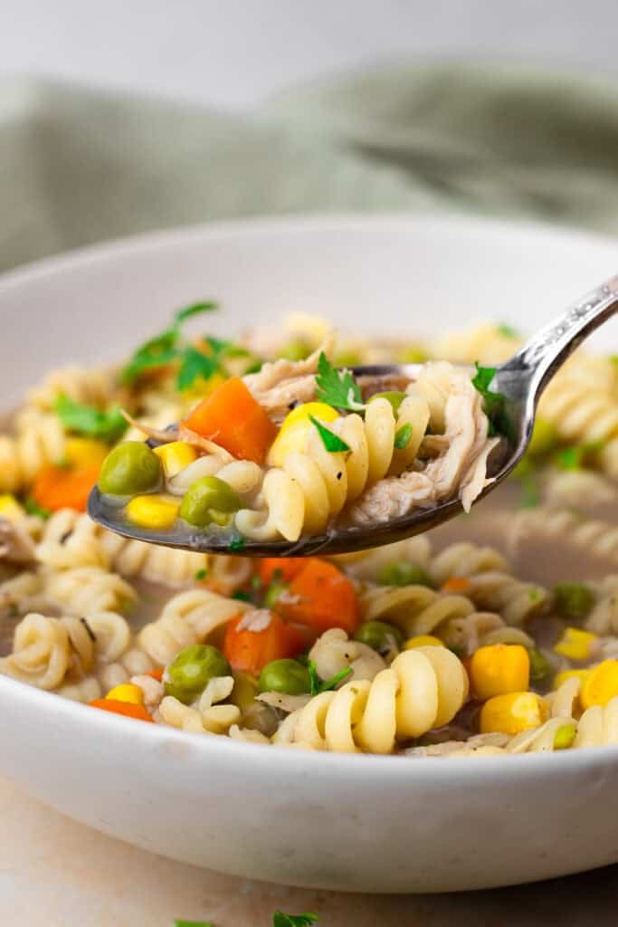 Side view image of spoon scooping up gluten free chicken noodle soup with corn, carrots, peas, and spiral noodles