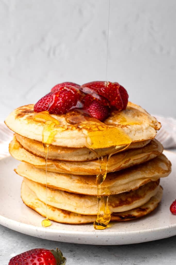 Gluten free pancakes in a stack with syrup being poured over it and strawberries