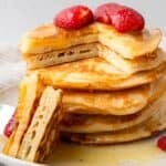 gluten free pancakes in a stack with a piece cut out and strawberries on top with maple syrup being poured on
