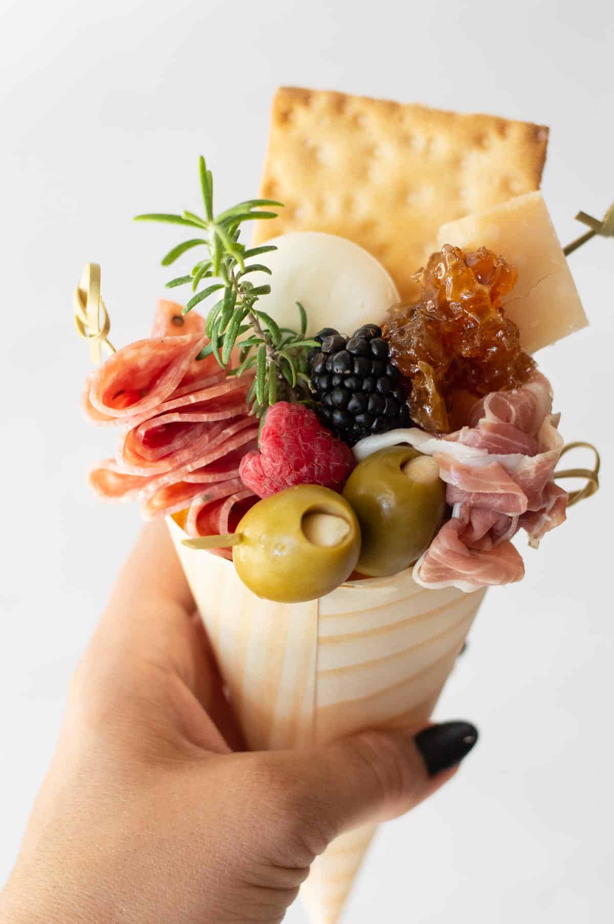 Charcuterie foods (meat, olives, blackberries, raspberries, cheese, crackers, and greenery) put into a brown cone.
