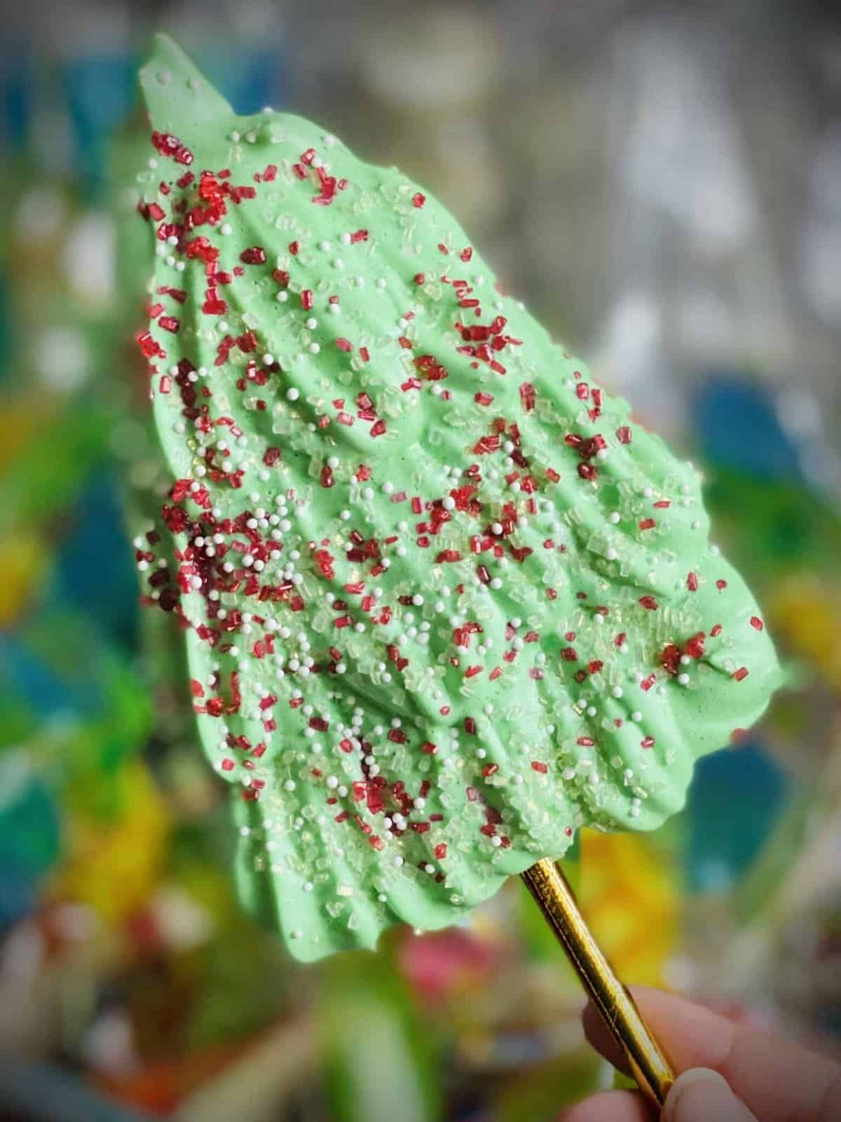 Vegan meringue cookies. It is a green Christmas tree on a gold stick. The tree is covered in red and white sprinkles.
