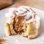 Single cinnamon roll sitting on a speckled plate, slathered in cream cheese frosting and a bite taken out to show the soft texture.