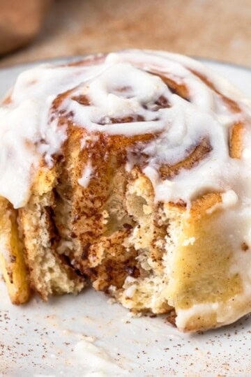 Single cinnamon roll sitting on a speckled plate, slathered in cream cheese frosting and a bite taken out to show the soft texture.