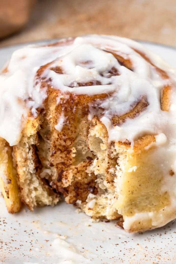 Single cinnamon roll sitting on a speckled plate, slathered in cream cheese frosting and a bite taken out to show the soft texture.