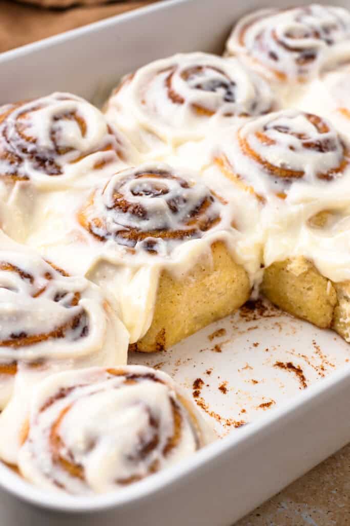 White baking dish filled with gluten-free cinnamon rolls in, slathered in cream cheese froster. One rolls has been removed from the pan leaving yummy cinnamon bits behind.