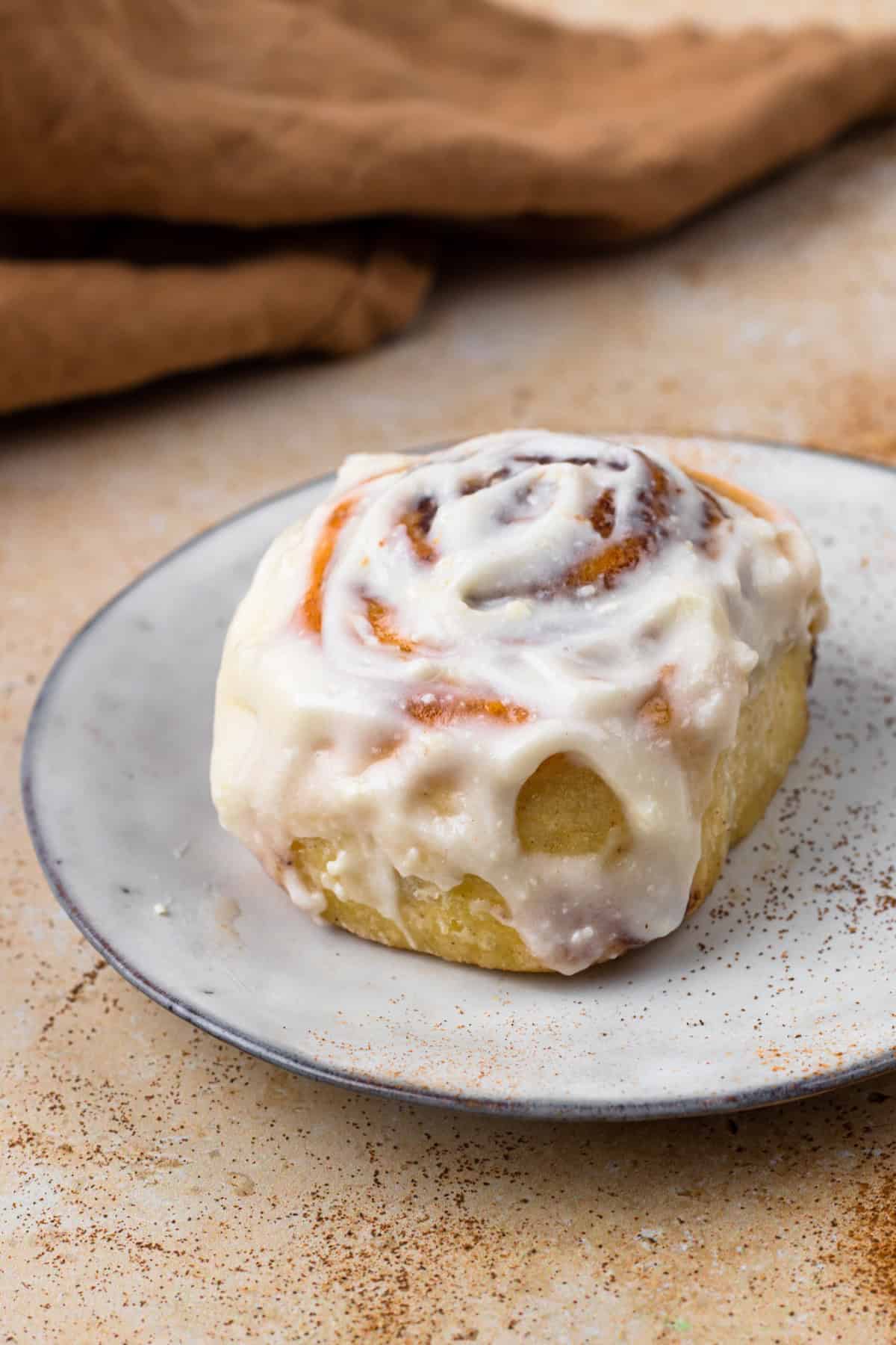 A single, frosting cinnamon roll sitting on top of a plate sprinkled with cinnamon.