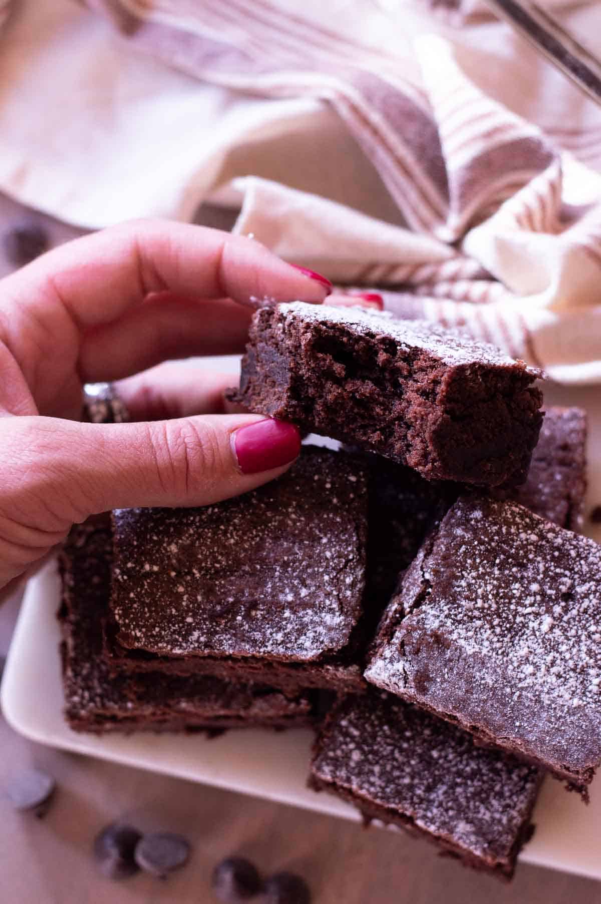 chewy gluten free brownies stacked on each other with powdered sugar and a bite taken out of one and a hand holding it