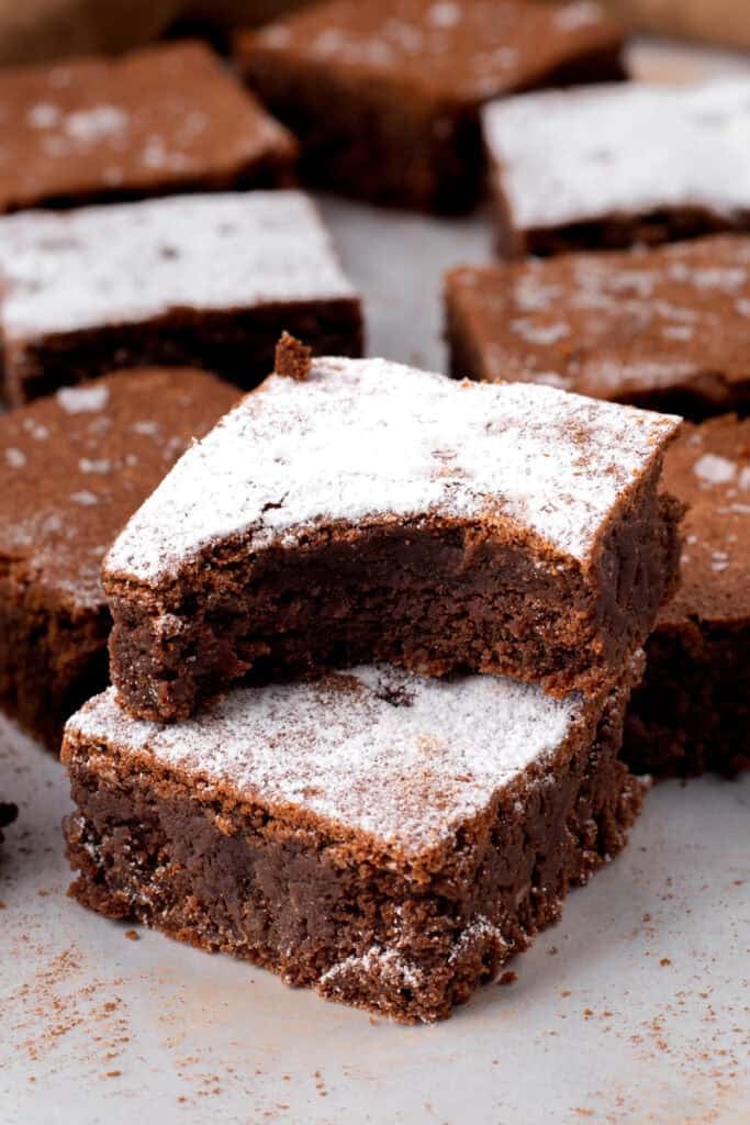 Side view of two gluten free brownies stacked on top of each other both topped with powdered sugar and one with bite taken out to show chewy texture