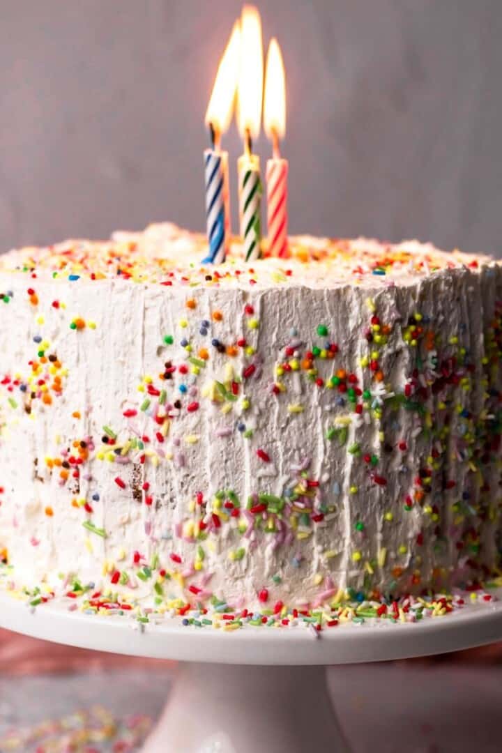 Gluten free birthday cake decorated with white frosting and sprinkles on a white cake plate with three lit candles on top.
