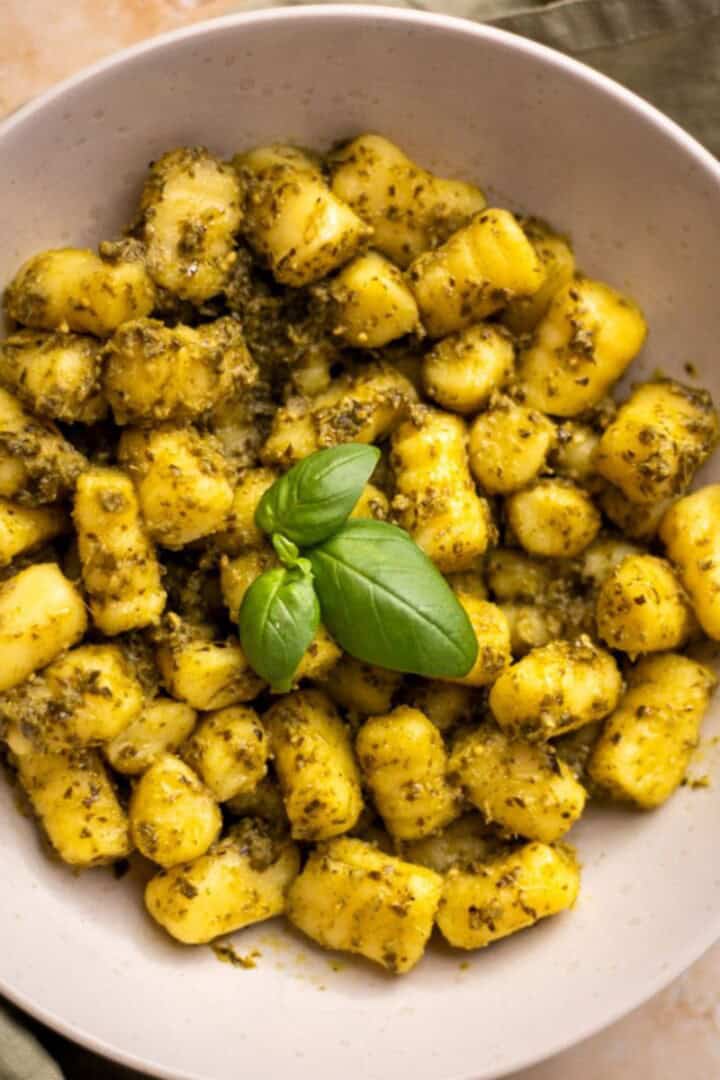 Seasoned gnocchi in a tan bowl with three basil leaves on top, the bowl is placed on a sage green cloth, more basil leaves are placed on the counter next to the bowl.