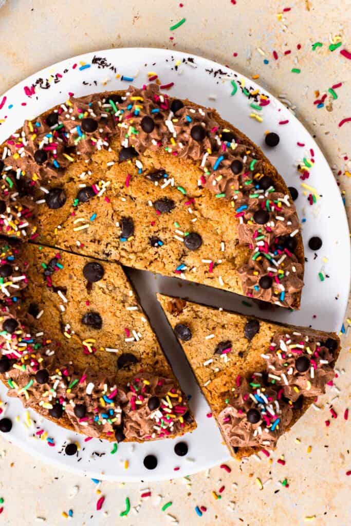 Top view of a gluten free cookie cake with a slice taken out, topped with chocolate buttercream on the outside, sprinkles and chocolate chips, sitting on a white plate, and surrounded by more sprinkles.