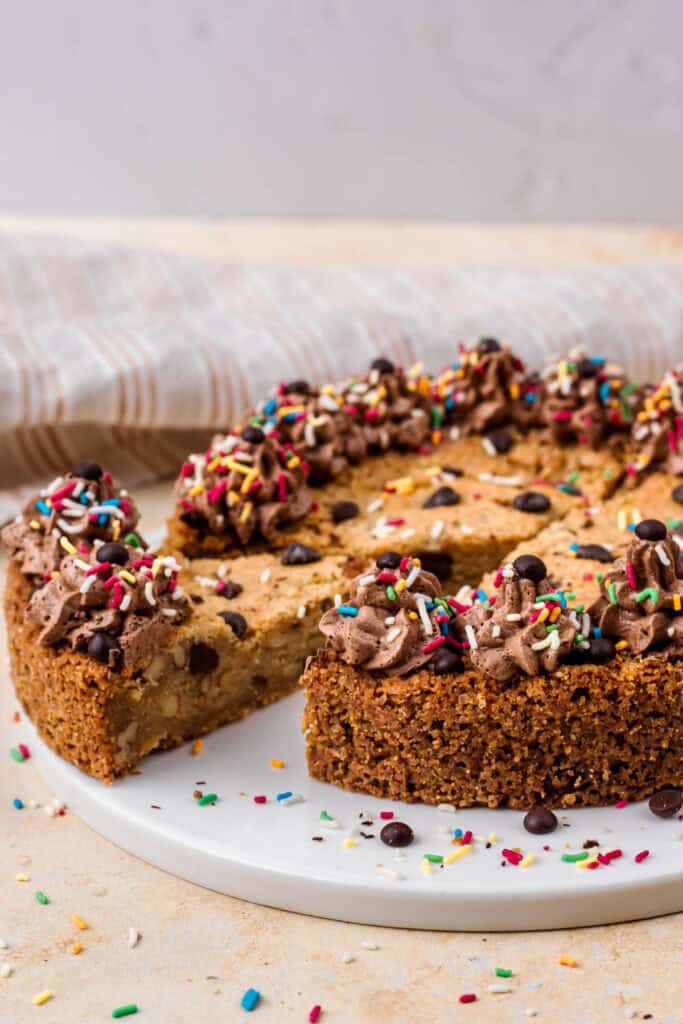 Side view of a gluten free cookie cake with a piece cut out, topped with chocolate buttercream on the outside, sprinkles and chocolate chips, sitting on a white plate, and surrounded by more sprinkles.