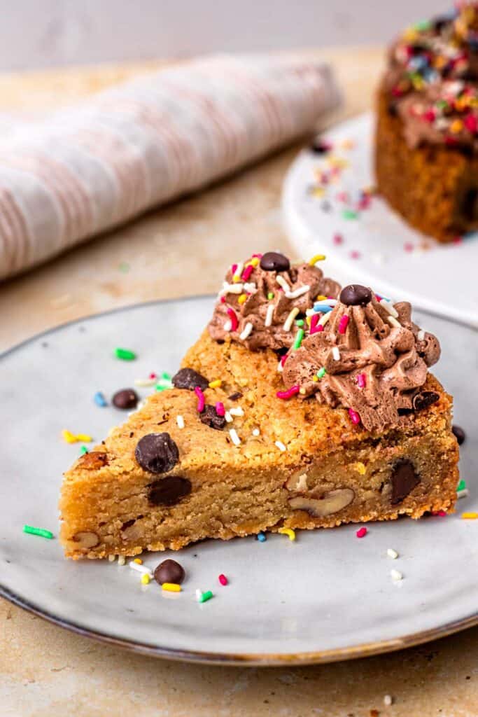Slice of a gluten free cookie cake topped with chocolate buttercream on the outside, sprinkles and chocolate chips, sitting on a white plate, and surrounded by more sprinkles.