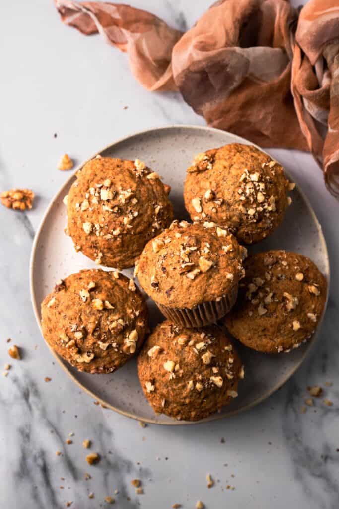 Six muffins on white plate on a white marble countertop.