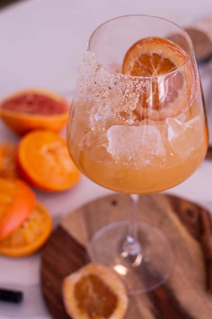 Glass of orang liquid with a candied orange slice, resting on a wooden cutting board near oranges that are blurred in the background.