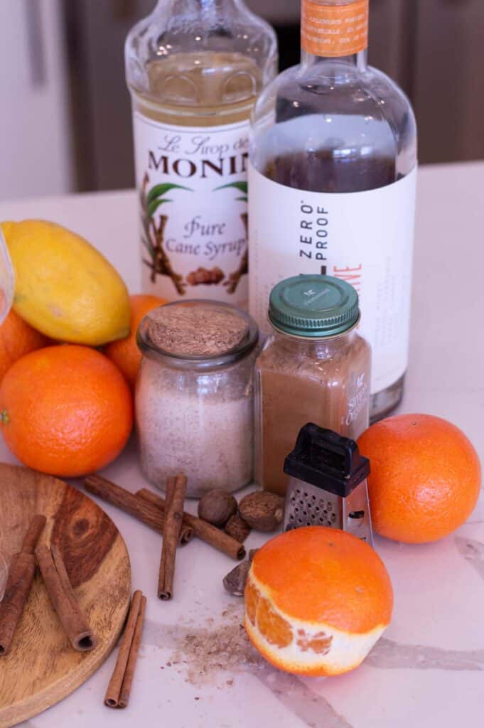 Ingredients for and Orange Mocktail, including cinnamon sticks, oranges, a lemon, a small grater, zero proof rum, simple syrup, and spices