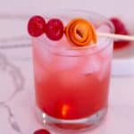 Glass filled with a Shirley Temple Mocktail with a stick of maraschino cherries and rolled orange peel balanced on top.