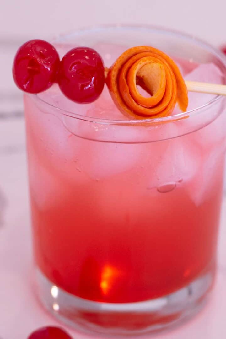 Glass filled with a Shirley Temple Mocktail with a stick of maraschino cherries and rolled orange peel balanced on top.