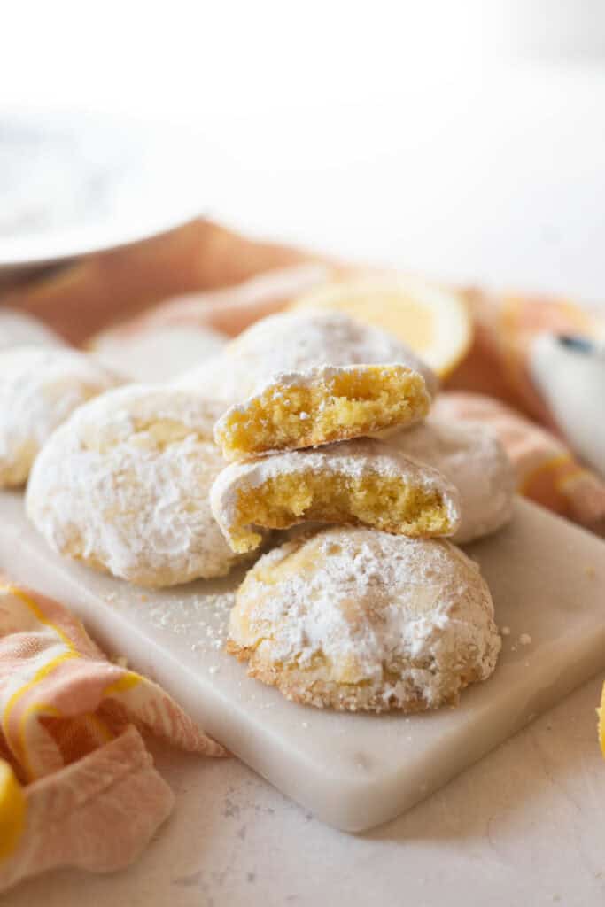Platter of gluten-free lemon crinkle cookies on top of a tea towel, the top cookie is broken in half to show the soft, chewy texture.