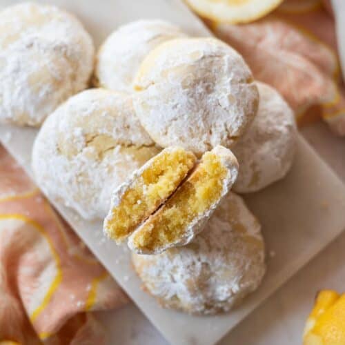 Square image of a gluten-free lemon crinkle cookie broken in half to show the chewy texture, resting on top of a platter of more cookies.